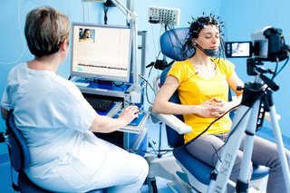 A person sitting on a chair, wearing a set of electrodes on their head while a health professional checks the EEG recording on a computer and a video recording is taken.