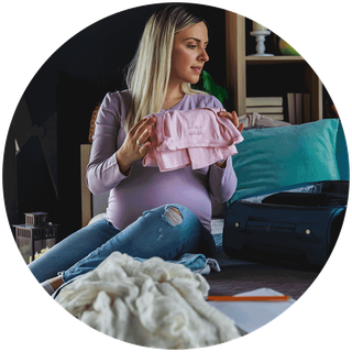 A pregnant mum to be is sitting on the bed, folding clothes ready to pack them into a small open suitcase.